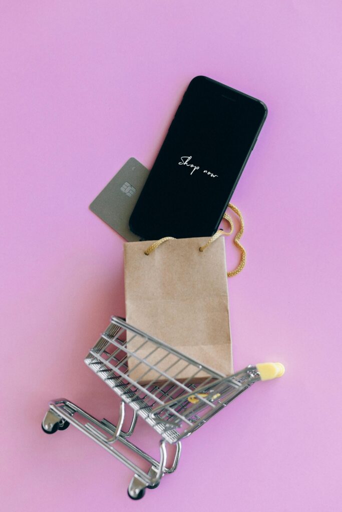 pexels-photo-6214450-6214450 A smartphone, credit card, and bag in a miniature shopping cart on a pink background.
